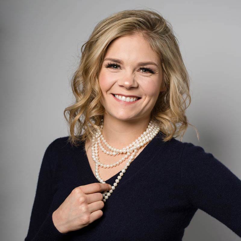 Headshot in studio of a blonde woman wearing pearls and a navy sweaterand smiling with a friendly smile and her hand on her hip.